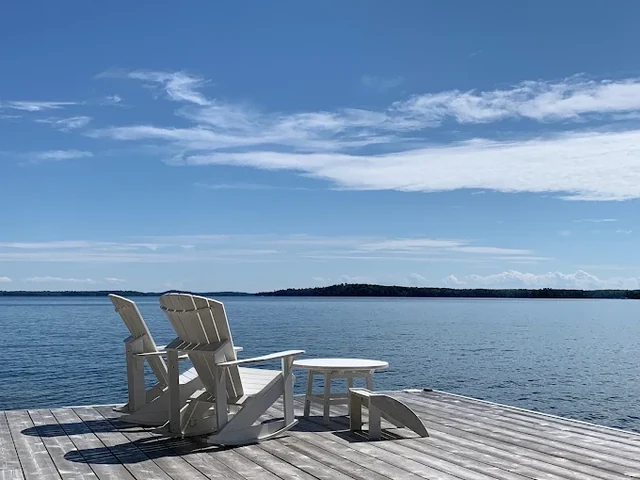Two white Adirondack chairs and a table on the sun dock at Sterling Point, looking out over the open water of Lake Joseph